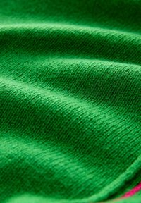 Vihreä neulottu kangas, jossa on pehmeä pinta, näkyvissä hienoja ompeleita sekä hienovaraisia sävy- ja kudontakuvion vaihteluita.