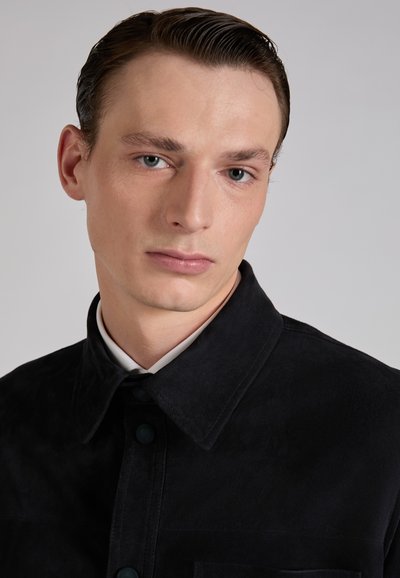 Młody mężczyzna z zaczesanymi do tyłu włosami, ubrany w czarną zapinaną na guziki kurtkę na białą koszulę, na jednolitym szarym tle.