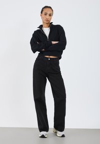 Sweat à zip noir en polaire avec un col haut, associé à un jean en denim noir taille haute. Des baskets de sport grises complètent le look.