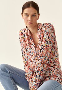 Blomsterbluse med en blanding af pink, orange og blå blomster på en lys baggrund, med V-udskæring og lange ærmer.