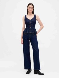 Donna che indossa un top peplum senza maniche di denim blu scuro con bottoni e jeans dritti abbinati, con stivaletti neri alla caviglia.
