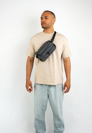 Sort taljerem med et strømlinet design, justerbar rem og detaljer i elastisk snor. Bæres tværs over kroppen over en beige skjorte og lyse jeans.