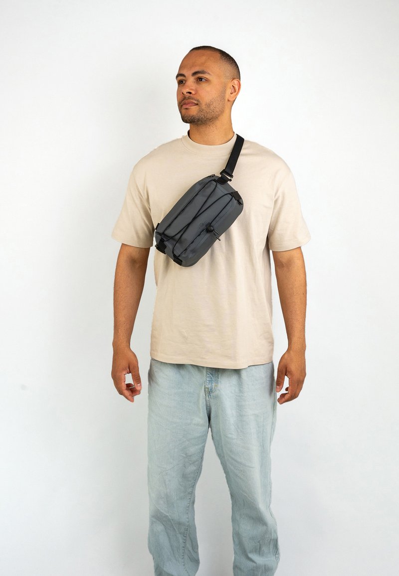 Borsa a vita nera dal design elegante, con tracolla regolabile e dettagli in corda elastica. Indossata a tracolla sopra una camicia beige e jeans chiari.