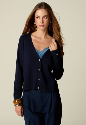 Jeune femme aux longs cheveux bruns porte un cardigan bleu marine sur un haut en dentelle bleu, un pantalon bleu marine, de grandes boucles d'oreilles et plusieurs bracelets.