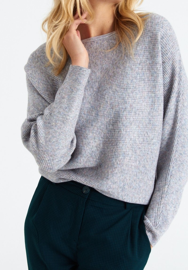 Pull gris clair à côtes avec une coupe décontractée, présentant des variations subtiles de couleur. Associé à un pantalon à carreaux vert foncé avec un bouton à la taille.