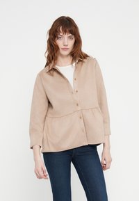 Light beige button-up shirt with a peplum design, long sleeves, and textured fabric. Paired with dark blue jeans. Simple and versatile style.