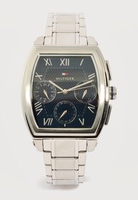 Ασημένιο μεταλλικό ρολόι χειρός με πρόσωπο σε ναυτικό μπλε, με ρωμαϊκούς αριθμούς, τρεις υποκαντράν για λειτουργίες 24 ωρών, ημέρας και ημερομηνίας, και το λογότυπο Hilfiger.