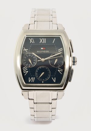 Ασημένιο μεταλλικό ρολόι χειρός με πρόσωπο σε ναυτικό μπλε, με ρωμαϊκούς αριθμούς, τρεις υποκαντράν για λειτουργίες 24 ωρών, ημέρας και ημερομηνίας, και το λογότυπο Hilfiger.