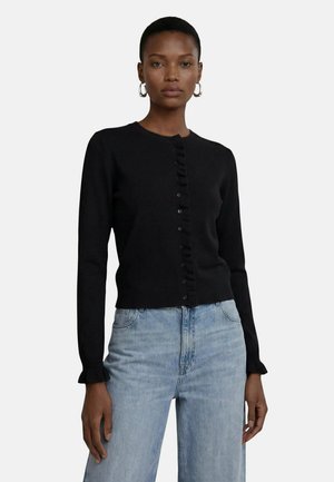 Femme portant un cardigan noir boutonné avec des détails à volants et un jean taille haute bleu clair, debout devant un fond blanc uni.