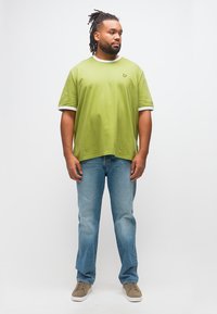 Άνδρας με κοτσιδάκια, ντυμένος με ανοιχτό πράσινο t-shirt, μπλε τζιν και μπεζ αθλητικά παπούτσια, στέκεται και κοιτάζει στο πλάι μπροστά σε απλό λευκό φόντο.