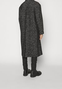 Černý kabát se vzorem rybí kosti s bílými akcenty, dlouhý, s texturovanou látkou a bočními rozparky. Nošený s úzkými černými kalhotami a pevnými botami.