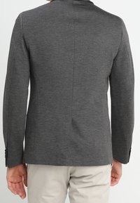 Uomo che indossa un blazer grigio scuro aderente e pantaloni beige chiaro, mostrato di spalle su uno sfondo semplice.