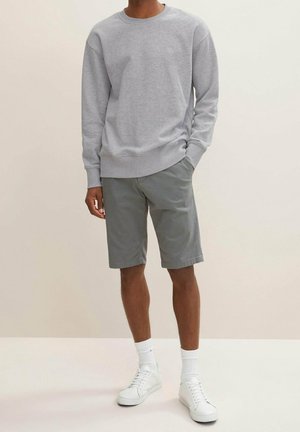 Mann trägt ein hellgraues Sweatshirt, graue kniehohe Shorts, weiße Sportsocken und weiße Turnschuhe und steht vor einem schlichten Hintergrund.