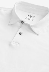 Polo de algodón blanco con cuello doblado, tapeta de dos botones y un pequeño detalle de bolsillo. La etiqueta interior indica la marca y la talla.