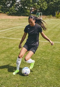 T-shirt de sport noir, shorts noirs, chaussettes blanches et crampons vert vif. Dribble d'un ballon de soccer multicolore sur un terrain en herbe.