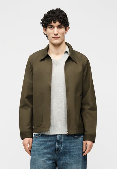 Jeune homme aux cheveux bouclés portant une veste zippée vert olive sur un pull gris clair et un jean bleu, debout devant un fond blanc uni.