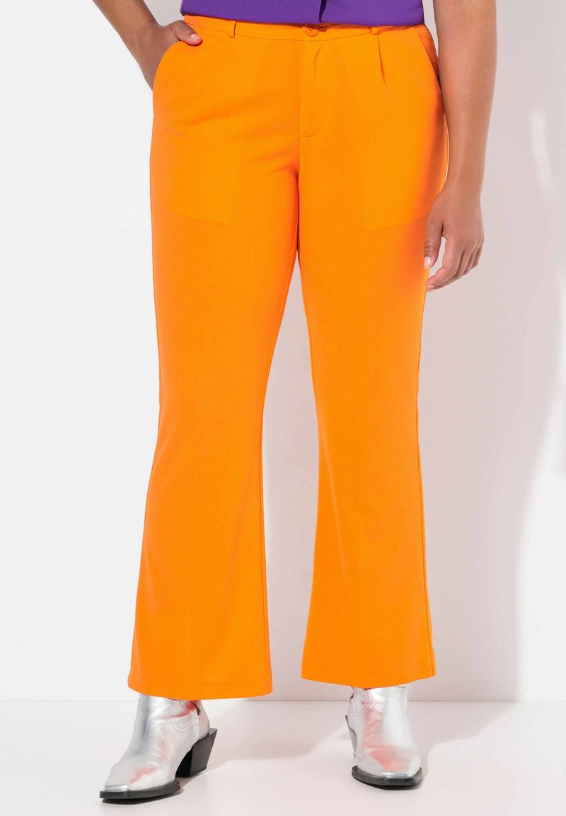 Ausgestellt geschnittene orangefarbene Hosen aus glattem Stoff, mit zwei vorderen Taschen und einem taillierten Bund. Getragen mit glänzenden silbernen Knöchelstiefeln.