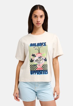 Žena má na sebe krémové tričko s farebným grafickým obrázkom vázy a kvetov s nápisom „Balance Opposites“ a svetlé rifľové šortky.