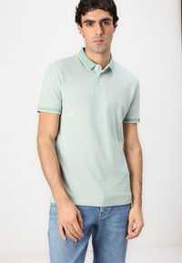 Lysegrønn polo med krage, med knappestolpe og kontrastkant på ermene. Brukt sammen med blå denimjeans.