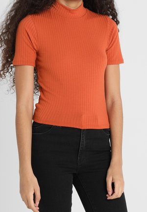 Vrouw draagt een nauwsluitende, korte mouw, geribbelde oranje coltrui en een hoog getailleerde zwarte jeans, staand tegen een effen achtergrond.