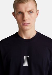 Mladý muž s krátkými blond vlasy, oblečený v černé košili s bílým nápisem "OUT" na hrudi, na neutrálním pozadí.