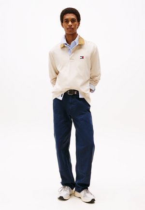 Polo beige a maniche lunghe con colletto in velluto a coste, pantaloni cargo blu navy e sneakers grigie, con un piccolo logo sul petto.