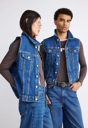 Kaks inimest, kes kannavad pruuni pika varrukaga särkide peal siniseid teksaveste ja siniseid teksapükse, seisavad ühetaustalise hele tausta ees.