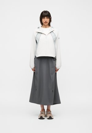Femme debout, vêtue d'une veste à capuche blanche, d'une longue jupe grise avec des cordons et de chaussures de sport beiges sur un fond blanc uni.