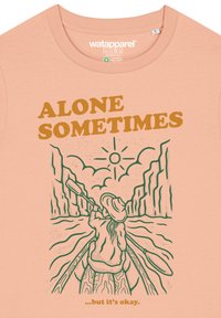 Perzikkleurig katoen T-shirt met groene lijntekening van een persoon die tussen kliffen kanoë, met de tekst "SOMS ALLEEN" erboven en een zon.