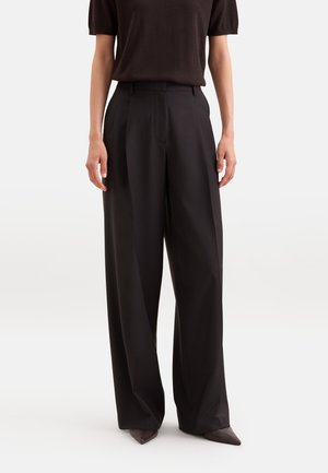 Personne portant un pantalon noir taille haute à jambes larges, associé à un haut noir à manches courtes et des chaussures à bouts pointus.