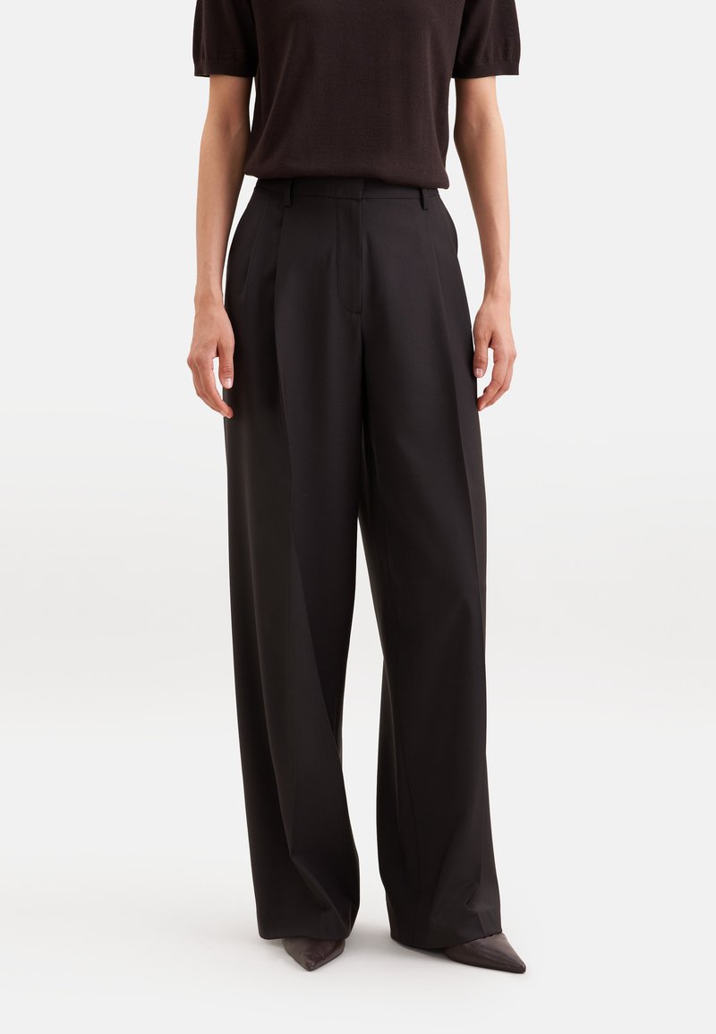 Personne portant un pantalon noir taille haute à jambes larges, associé à un haut noir à manches courtes et des chaussures à bouts pointus.