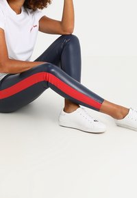 Legginsy z wysokim stanem, w kolorze granatowym z ekoskóry, z czerwonym paskiem po boku, zestawione z białym t-shirtem i białymi trampkami. Gładka tekstura, elegancki design.