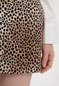 Gros plan d’une main à côté d’une jupe à imprimé léopard avec des taches noires sur fond beige et une manche de chemise blanche.