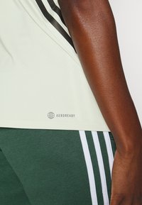 Gros plan d'une personne portant un maillot de sport de couleur claire avec le logo "AEROREADY" et un short de sport vert avec trois bandes blanches.