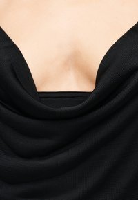 Czarna prążkowana tkanina delikatnie opada na skórę, odsłaniając głęboki dekolt z widocznym obojczykiem i górną częścią klatki piersiowej.