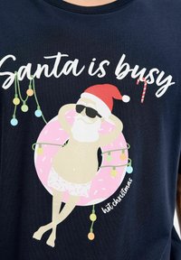 Marineblauwe T-shirt met een kerstman in een korte broek en zonnebril, die op een roze donut met glazuur ligt, omringd door kleurrijke lampjes en tekst.