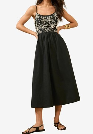 Femme portant une robe midi noire à fines bretelles avec une broderie florale crème sur le corsage, associée à des sandales noires et des bracelets dorés.