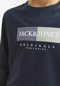 Tamsiai mėlyna medvilninė ilgomis rankovėmis marškinėliai su grafiniu piešiniu, kuriame pavaizduoti užrašai „JACK&JONES“ ir „ORIGINALS WORLDWIDE“ balta ir pilka spalvomis.