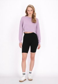 Afgeknotte paarse sweatshirt met lange mouwen, gecombineerd met hoge zwarte fietsbroek. Het model draagt witte sneakers en gestreepte sokken.