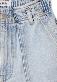 Jeans in denim azzurro chiaro con una texture liscia, caratterizzati da cuciture visibili, un bottone robusto e un cinturino con arricciature elastiche per maggiore flessibilità.