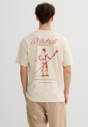 Hombre con camiseta crema que lleva una ilustración roja de un remero sosteniendo un remo y el texto "Whitehall Rowing Society" en la parte trasera.