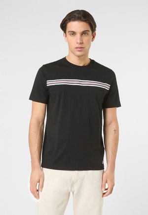 Ung mand med kort mørkt hår iført en sort T-shirt med hvide, røde og blå vandrette striber, stående foran en ensartet lys baggrund.