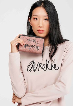 Cartera acolchada rosa con letras decorativas en negro y rhinestones, sostenida por una persona que lleva una camiseta rosa claro con un logo negro contrastante.