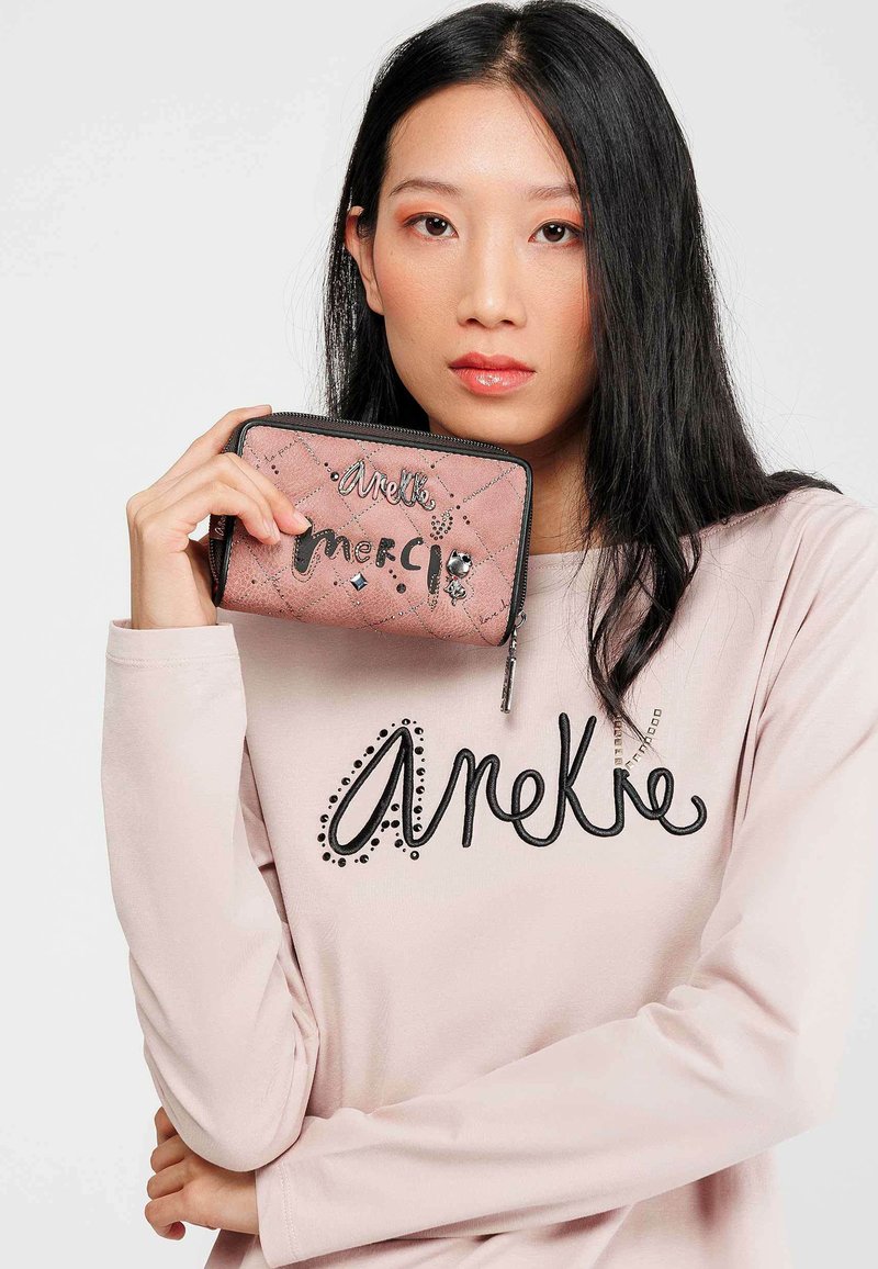 Cartera acolchada rosa con letras decorativas en negro y rhinestones, sostenida por una persona que lleva una camiseta rosa claro con un logo negro contrastante.