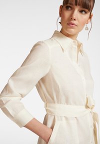 Abito camicia color crema con colletto, maniche lunghe, chiusura a bottoni e cintura annodata in vita. Tessuto leggero e testurizzato con tasche.