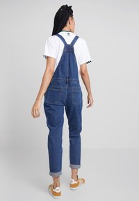 Nainen, jolla on pitkät letit, pukeutuneena sinisiin farkkuhaalareihin, valkoiseen Adidas T-paitaan ja sinappikeltaisiin Adidas-tennareihin, seisoo selkä kameraan päin.