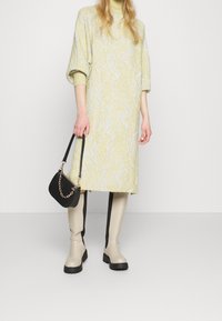 Knälång gul stickad klänning med vita vågmönster, i kombination med beige knähöga stövlar och en liten svart handväska med kedja.