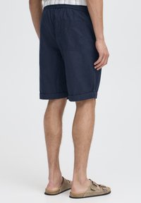 Pantaloni scurți bleumarin din material ușor, cu buzunar pe spate, tiv răsucit și talie elastică. Purtate cu sandale maro tip slide.