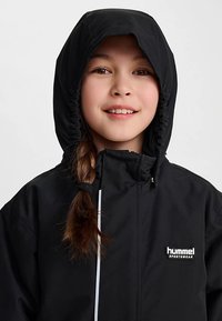 Sort vandtæt jakke med hætte, der har lynlås og snøre. Bemærkelsesværdigt "hummel"-logo på brystet. Glat tekstur.