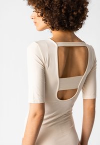 Body beige con maniche corte, caratterizzato da un design con schiena scoperta e un taglio rettangolare. Materiale liscio e elasticizzato con una vestibilità aderente.